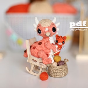 May include: A crocheted pink dinosaur with white spots sits on a rocking chair holding a crocheted orange tiger. The dinosaur is holding a knitting needle and a basket of yarn balls.