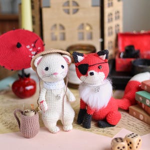 May include: Two crocheted stuffed animals, a white ferret wearing a straw hat and a red fox wearing an eye patch, sitting on a pink surface with two brown dice.