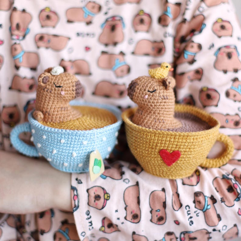 May include: Two crocheted capybara figurines are sitting in teacups. The capybara on the left is in a blue teacup with white polka dots. The capybara on the right is in a yellow teacup with a red heart. Both capybaras are brown and have closed eyes.