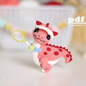 May include: A pink crocheted dinosaur holding a green tennis ball and a green and white tennis racket. The dinosaur is wearing a white crocheted hat with a red brim. The dinosaur is standing on a white surface.  pdf LoopyPattern