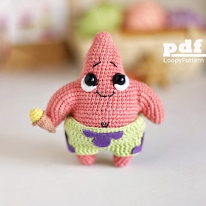 Könnte beinhalten: Gehäkelte rosa Seesternfigur mit großen schwarzen und weißen Augen, die ein gelbes Eis halten. Die Figur trägt hellgrüne Shorts mit lila Akzenten. Der Text "pdf LoopyPattern" ist sichtbar.