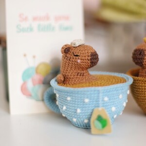 May include: A brown crocheted capybara with closed eyes is sitting in a blue teacup with white polka dots. The teacup is filled with brown yarn and has a tea bag tag attached to the side. The text "So much you, Such little time" is visible on a card in the background.