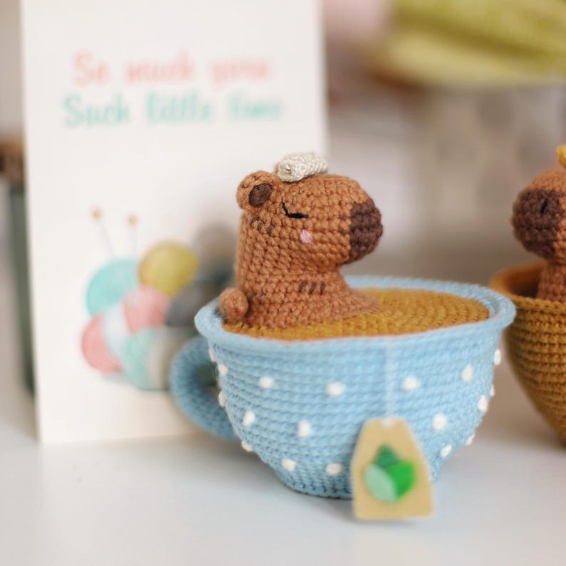 May include: A brown crocheted capybara with closed eyes is sitting in a blue teacup with white polka dots. The teacup is filled with brown yarn and has a tea bag tag attached to the side. The text "So much you, Such little time" is visible on a card in the background.