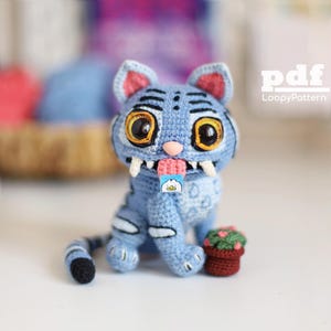 Puede incluir: Una figura de tigre tejida a ganchillo en color azul con grandes ojos ámbar, colmillos blancos y una lengua rosa que sostiene una pequeña etiqueta cuadrada con un gráfico de pájaro blanco. El tigre tiene rayas azul oscuro y una cola larga. Texto pdf LoopyPattern.
