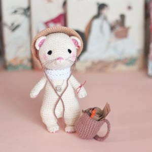 May include: A white crocheted ferret wearing a brown hat and a white scarf, holding a brown crocheted mug with a rolled up piece of paper inside.