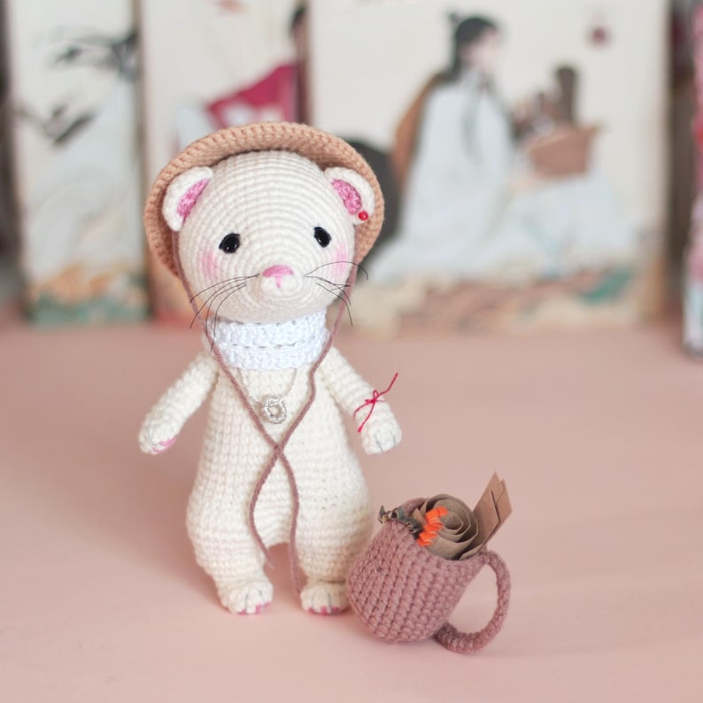 May include: A white crocheted ferret wearing a brown hat and a white scarf, holding a brown crocheted mug with a rolled up piece of paper inside.