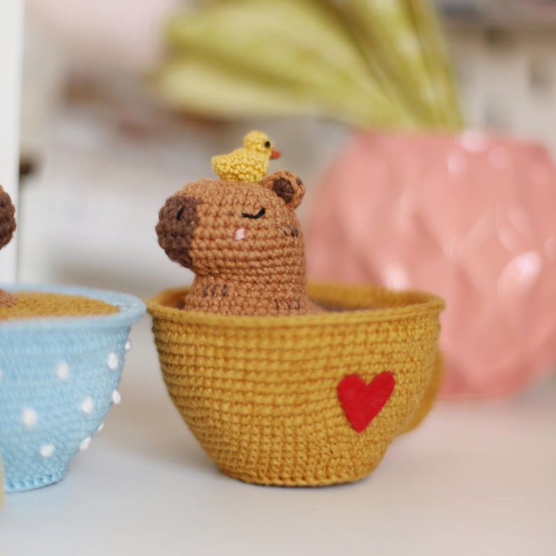 May include: A brown crocheted capybara with a yellow crocheted duck on its head sits in a yellow crocheted teacup with a red heart on the side.