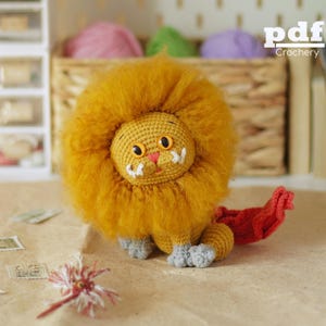 May include: A crocheted lion toy with a large, fuzzy mustard-yellow mane, orange eyes, a pink nose, and white embroidered whiskers. The body is mustard-yellow with light gray paws and a long, textured red tail extending to the right side.