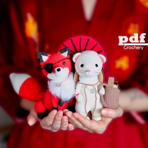 Puede incluir: Dos juguetes amigurumi de crochet, un zorro rojo con un parche en el ojo y un hurón blanco con un sombrero rojo, se sostienen en las palmas de las manos de una persona. El zorro tiene una cola blanca y esponjosa y un collar blanco con un dije plateado. El hurón tiene una bolsa de crochet marrón en su hombro. El fondo es una tela roja.
