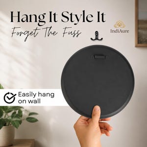 May include: A round black wall-mounted object with a flat back and a metal hanging bracket. The text reads Hang It Style It Forget The Fuss and Easily hang on wall. A small black double hook is positioned above the round object.