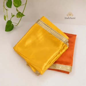 Op de afbeelding: Een gevouwen felgeel stuk stof dat over een gevouwen oranje stuk stof ligt, beide voorzien van een metallic gouden geborduurde rand. De merknaam IndiAure staat in de rechterbovenhoek boven de stoffen gedrukt.