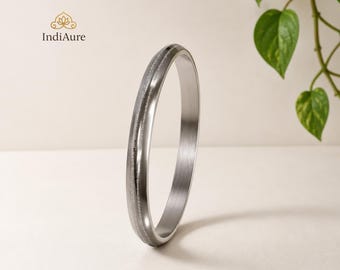 May include: A silver-toned metal bangle bracelet with a smooth, polished finish and a thin, textured central groove running along the circumference. The circular band features a clean, minimalist design with a reflective surface and rounded edges.