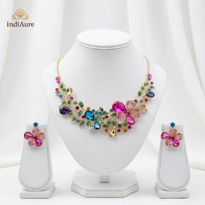 May include: A floral necklace and matching earrings set. The jewelry features colorful, faceted gemstones in shades of pink, blue, purple, and yellow, set in a gold-tone metal. The necklace and earrings are displayed on white jewelry stands.