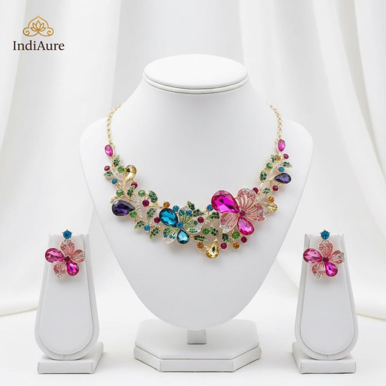 May include: A floral necklace and matching earrings set. The jewelry features colorful, faceted gemstones in shades of pink, blue, purple, and yellow, set in a gold-tone metal. The necklace and earrings are displayed on white jewelry stands.