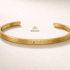 May include: A gold-colored cuff bracelet with engraved text and a small figure. The bracelet has a smooth, polished finish and a simple, open design. The text appears to be in a foreign language. The brand name "IndiAure" is also visible.