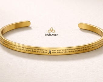May include: A gold-colored cuff bracelet with engraved text and a small figure. The bracelet has a smooth, polished finish and a simple, open design. The text appears to be in a foreign language. The brand name "IndiAure" is also visible.