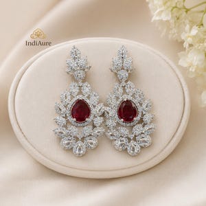 Peut inclure: Une paire de boucles d'oreilles argentées ornées, avec un centre en pierre précieuse rouge foncé en forme de goutte d'eau. Les boucles d'oreilles présentent un motif floral avec de nombreux petits cristaux transparents. Elles sont présentées sur une surface crème.