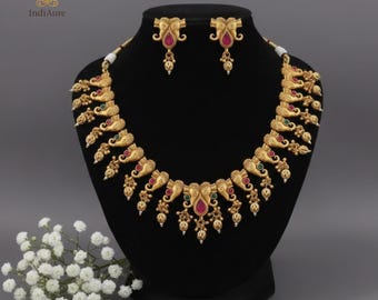 May include: A gold-toned necklace and matching earrings set. The necklace features an intricate design with elephant motifs, red and green accents, and dangling pendants. The earrings mirror the design with red gemstones.