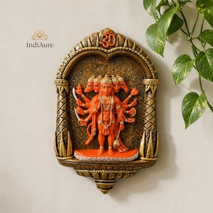 May include: A detailed orange-colored wall hanging depicting a multi-faced deity with multiple arms. The artwork is set within an arched frame with gold and brown accents, and the word "Om" is at the top. The piece is from the brand "IndiAure".