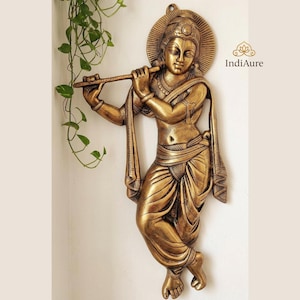 May include: A golden metal wall sculpture of a figure playing a flute. The figure wears traditional garments and has a halo. The sculpture is detailed and mounted on a wall. The IndiAure logo is visible.