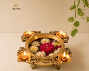 Puede incluir: Un cuenco redondo de latón con bordes de volutas ornamentadas y cuatro portavelas integrados, cada uno con una mecha encendida. El cuenco está lleno de agua y flores blancas y rosa intenso flotantes. El texto IndiAure es visible arriba a la izquierda.