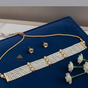 Puede incluir: Un collar gargantilla de perlas y un conjunto de pendientes a juego. El collar presenta múltiples hilos de perlas blancas con detalles dorados y gemas transparentes. El conjunto de joyas se exhibe sobre una superficie azul.