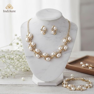 Op de afbeelding: Een goudkleurige sieradenset met een ketting, oorbellen en armband, allemaal versierd met imitatieparels en kleine heldere kristallen. De ketting en armband hebben een bloemmotief. De sieraden worden getoond op een witte marmeren buste.