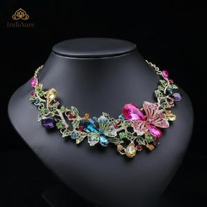 May include: A floral statement necklace with colorful gemstones. The necklace features a gold-tone chain and is adorned with pink, blue, green, yellow, and purple crystals arranged in a flower and leaf design. The necklace is displayed on a black mannequin.
