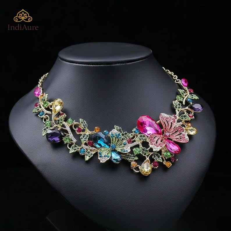 May include: A floral statement necklace with colorful gemstones. The necklace features a gold-tone chain and is adorned with pink, blue, green, yellow, and purple crystals arranged in a flower and leaf design. The necklace is displayed on a black mannequin.