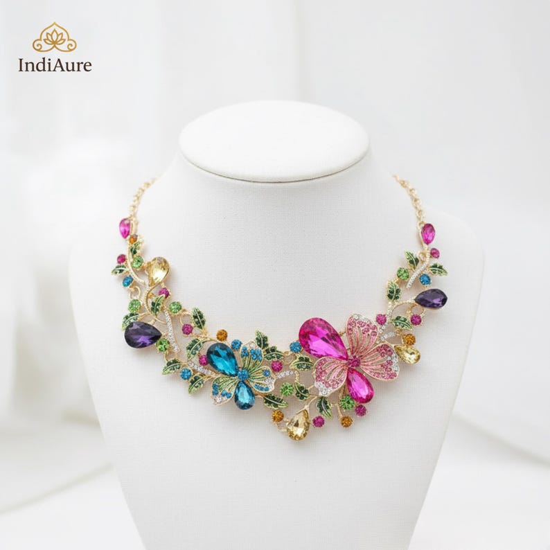 May include: A gold-toned necklace with a floral design, featuring large, colorful gemstones in shades of pink, blue, yellow, and purple. The necklace is displayed on a white mannequin bust. The brand name "IndiAure" is visible in the upper left corner.