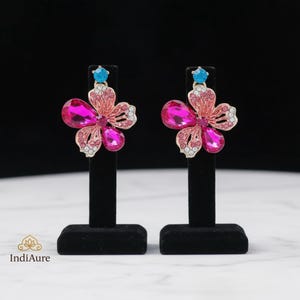 May include: A pair of flower-shaped earrings. Each earring features a pink and magenta crystal flower design with a small blue crystal at the top. The earrings are displayed on black stands against a black background.