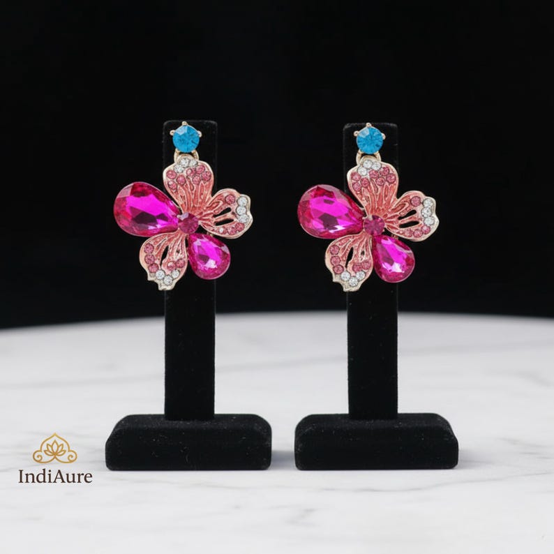 May include: A pair of flower-shaped earrings. Each earring features a pink and magenta crystal flower design with a small blue crystal at the top. The earrings are displayed on black stands against a black background.