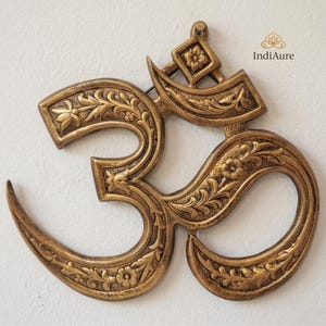 Könnte beinhalten: Ein detailliertes Om-Symbol aus Messing zum Aufhängen an der Wand. Das Symbol ist aufwendig mit floralen Mustern und Designs geschnitzt. Das goldfarbene Metallstück ist an einer weißen Wand befestigt. Es ist ein dekorativer Artikel.