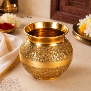 May include: A polished brass pot featuring a wide, flared rim and a rounded body. The exterior surface is decorated with intricate, embossed floral patterns and scrolling vine motifs, separated by a smooth, plain horizontal band around the center.