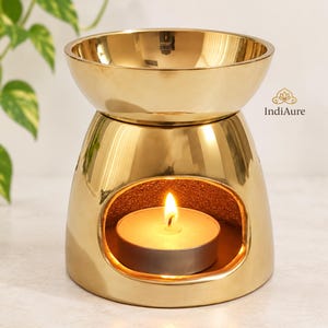 May include: A polished gold-colored metal oil burner with a wide bowl at the top and a circular opening at the base containing a lit white tea light candle. The brand name IndiAure is printed in small black text on the right side of the object.
