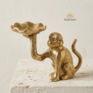 May include: A gold-colored brass monkey figurine holding a scalloped, bowl-shaped tray above its head. The monkey is depicted in a seated position with a curved tail. The text IndiAure is visible in the upper right corner of the image.