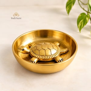Peut inclure: Un bol rond en métal doré poli avec une figurine de tortue en trois dimensions fixée au centre. La tortue présente une carapace segmentée détaillée et des membres texturés. Le nom de marque IndiAure est imprimé dans le coin supérieur gauche.