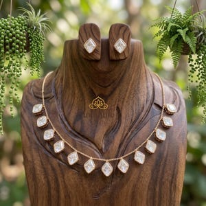 Könnte beinhalten: Ein goldfarbenes Halsketten- und Ohrring-Set, das auf einer Holzbüste präsentiert wird. Die Halskette besteht aus einer Reihe quadratischer, klarer Edelsteine. Die Ohrringe haben einen einzelnen quadratischen Edelstein. Der Hintergrund umfasst Hängepflanzen.