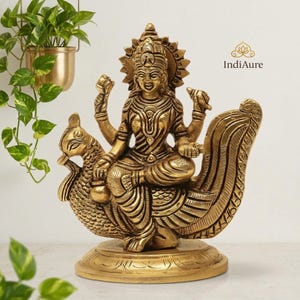 May include: A gold-colored statue of a deity seated on a peacock. The statue has intricate details, including the deity's jewelry and the peacock's feathers. The statue is on a round base and is likely made of brass.