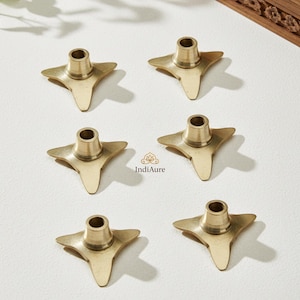 May include: Six brass-colored star-shaped candle holders. Each holder has a central cylindrical cup for the candle and three pointed star-shaped legs. The holders are arranged on a white surface.