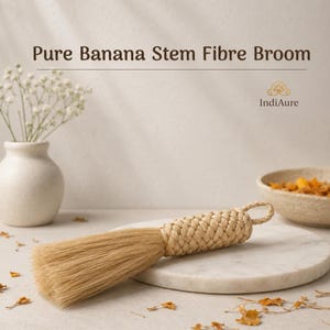 Op de afbeelding: Een bezem gemaakt van natuurlijke bananenstengelvezels, voorzien van een strak gevlochten handvat met een lus aan het uiteinde om op te hangen. De tekst Pure Banana Stem Fibre Broom en de merknaam IndiAure zijn bovenaan zichtbaar.