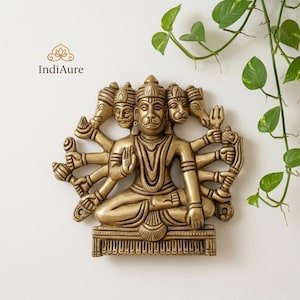 May include: A gold-coloured wall hanging of a multi-faced deity with multiple arms, seated on a decorative base. The sculpture has intricate details and is set against a white background with green foliage.