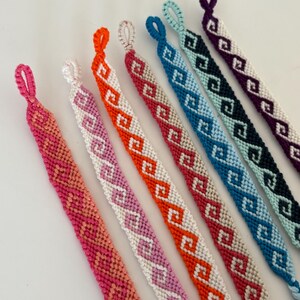 Könnte beinhalten: Eine Sammlung handgefertigter Freundschaftsarmbänder in verschiedenen Farben. Jedes Armband zeigt ein sich wiederholendes geometrisches Muster in Rosa-, Orange-, Weiß-, Blau- und Lilatönen. Die Armbänder werden auf einer weißen Oberfläche präsentiert.