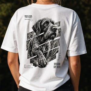 May include: A white t-shirt featuring a black and white graphic on the back of a wirehaired pointing dog in tall grass. The text reads BUILT FOR THE POINT, GUN DOGS, GRIFFON DIVISION, GP-1874, and includes coordinates and a small scale graphic.