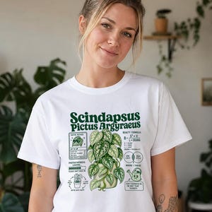 以下が含まれることがあります： Scindapsus Pictus Argyraeusの植物のダークグリーンのグラフィックがプリントされた白いTシャツ。デザインにはテキスト、植物のイラスト、光合成の読み込みバー、美容の公式、そして「Don't overwater me, I'm not that thirsty.」というテキストが書かれたじょうろのアイコンが含まれています。
