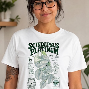 以下が含まれることがあります： Scindapsus Platinumの植物が描かれたダークグリーンのグラフィック入りホワイトTシャツ。デザインにはテキスト、周期表の要素ボックス、ケアレベルゲージ、ベン図、そしてHeart Stealerというテキストが書かれた指名手配ポスターのイラストが含まれています。