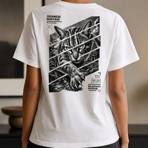 Pode incluir: Uma t-shirt branca com um gráfico a preto e branco nas costas. O design mostra um gato javanês com o texto JAVANESE HERITAGE, WILD ELEGANCE, SPEC. JV-01 FELIS JAVANENSIS e MAJAPAHIT DIVISION, inserido em painéis diagonais cortados.