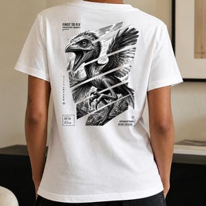 May include: A white t-shirt featuring a black and white graphic on the back of an Archaeopteryx dinosaur with wings spread. The design includes text reading FIRST TO FLY, PREHISTORIC ANIMALS, SPECIMEN ID ARC-150, LENGTH 0.5-0.6 M, and ARCHAEOPTERYX AVIANS DIVISION.