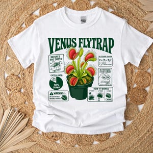Könnte beinhalten: Ein weißes T-Shirt mit einer Grafik einer Venusfliegenfalle in einem grünen Topf. Das Design enthält Text wie VENUS FLYTRAP, NATURE'S BUG ZAPPER, CATCH RATE: 90% DRAMA und ein Diagramm HOW IT WORKS, das die schließende Pflanze zeigt.