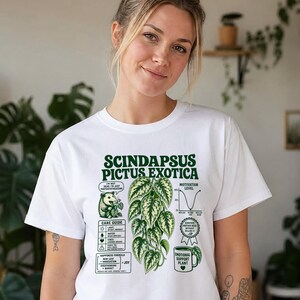 以下が含まれることがあります： Scindapsus Pictus Exoticaの植物の緑色のグラフィックがプリントされた白いTシャツ。デザインにはテキスト、ケアガイド、モチベーションレベルのグラフ、オポッサムのイラスト、Emotional Support Plantと書かれたマグカップが含まれています。